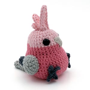 May include: A handmade crochet bird in shades of pink, gray, and black. The bird has a rounded body, a crest, and small feet. The eyes are black. The bird is sitting on a white surface.