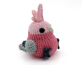 Op de afbeelding: Een handgemaakte gehaakte vogel in roze, grijs en zwart. De vogel heeft een afgerond lichaam, een kuif en kleine voetjes. De ogen zijn zwart. De vogel zit op een witte ondergrond.