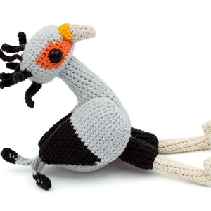 Può includere: Un giocattolo di peluche a forma di uccello lavorato a uncinetto con filato grigio, nero e bianco. L'uccello ha un collo lungo, un piccolo becco e grandi occhi arancioni. L'uccello è seduto sulla pancia con le gambe distese.