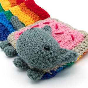 K&ouml;nnte beinhalten: Ein geh&auml;keltes Regenbogen- und graues Katzenspielzeug. Die Katze liegt auf der Seite mit dem Kopf auf dem Regenbogen.
