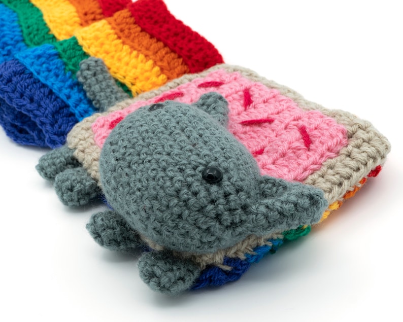 K&ouml;nnte beinhalten: Ein geh&auml;keltes Regenbogen- und graues Katzenspielzeug. Die Katze liegt auf der Seite mit dem Kopf auf dem Regenbogen.