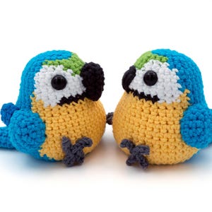 May include: Two handmade crochet parrots. Each bird has a blue head with green accents, white face, black beak, and yellow body. The parrots are positioned side-by-side, showcasing their detailed craftsmanship and vibrant colors.