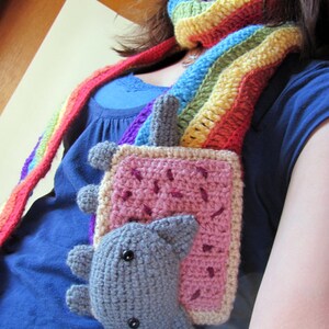 K&ouml;nnte beinhalten: Eine Frau tr&auml;gt einen Regenbogen-H&auml;kel-Schal mit einem geh&auml;kelten Katzen-Toast-Anh&auml;nger. Der Anh&auml;nger ist rosa, grau und braun.