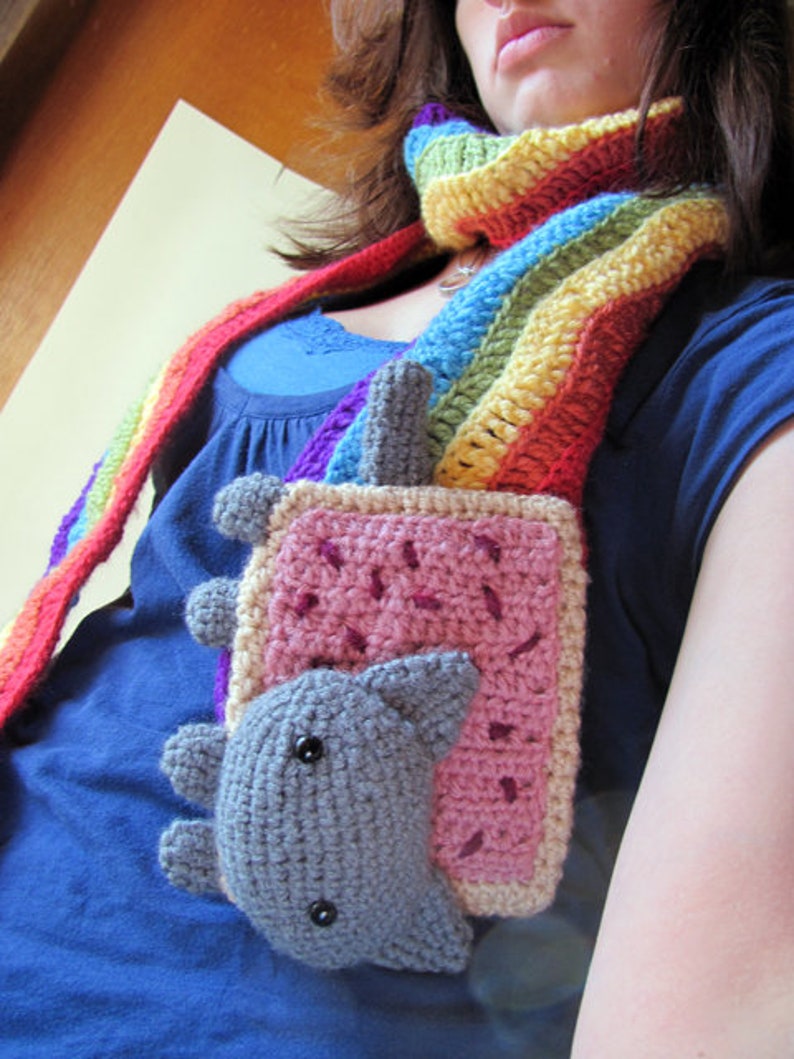 K&ouml;nnte beinhalten: Eine Frau tr&auml;gt einen Regenbogen-H&auml;kel-Schal mit einem geh&auml;kelten Katzen-Toast-Anh&auml;nger. Der Anh&auml;nger ist rosa, grau und braun.