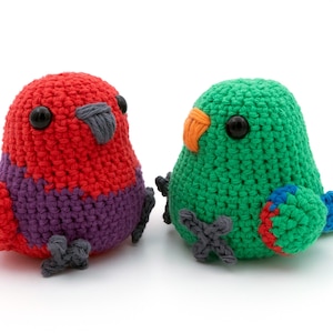 May include: Two handmade crochet birds. One is red and purple, the other is green with an orange beak. Both have black eyes and grey feet. The birds are side by side on a white surface.