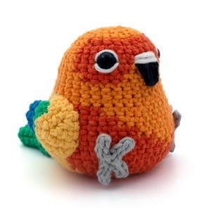 May include: A small, round, crocheted bird figurine featuring a vibrant orange and red body, yellow wings, and a green tail. The bird has large black plastic eyes, a black and white beak, and light gray crocheted feet attached to the front.