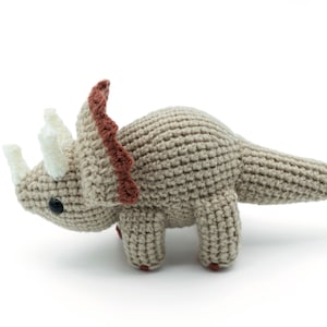 May include: A crocheted stuffed triceratops toy with a brown body, white horns, and a brown frill. The toy is made with a textured stitch and has a cute, cartoonish appearance.