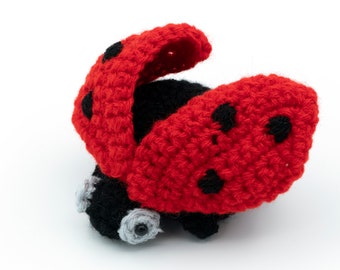 Puede incluir: Un juguete de mariquita de crochet con alas rojas y manchas negras. La mariquita tiene ojos negros y un cuerpo negro.