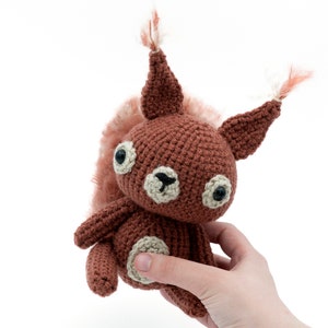 May include: A crocheted squirrel plush toy with brown fur, a fluffy tail, and black eyes. The toy is being held in a hand.
