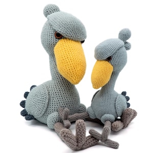 May include: Two crocheted stuffed birds, one larger than the other, both with gray bodies, yellow beaks, and gray legs. The birds are sitting on a white background.