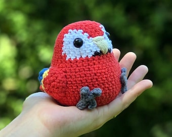 May include: A handmade, red crochet parrot with white and black accents around the eyes and a tan beak. The bird has gray feet and is perched on a hand. The background is a blurred green.