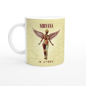 Peut inclure: Mug en céramique blanche avec l'illustration de l'album "In Utero" de Nirvana. Le design comprend le nom du groupe en bordeaux, une figure ailée et le titre de l'album. Le fond a un aspect vintage et texturé.