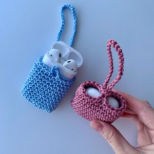May include: Two crochet AirPod cases, one blue and one pink, with matching braided straps. The blue case holds white AirPods. The pink case is held by a hand. The cases are made of a textured, woven material.