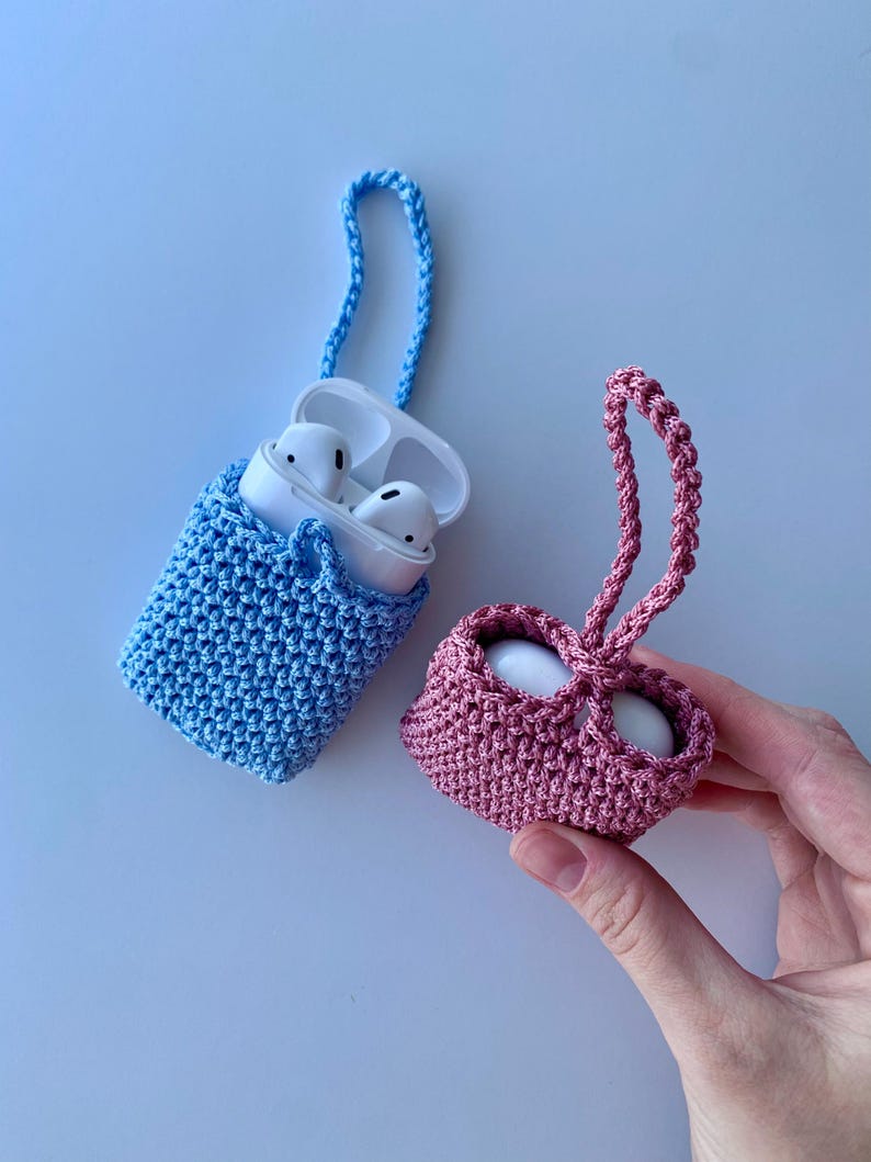 May include: Two crochet AirPod cases, one blue and one pink, with matching braided straps. The blue case holds white AirPods. The pink case is held by a hand. The cases are made of a textured, woven material.
