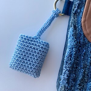 May include: A light blue, crocheted square pouch with a matching strap, attached to a silver metal ring. The pouch is connected to a denim bag with a brown leather accent.