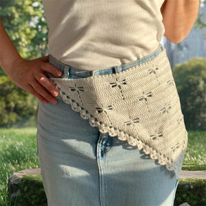 K&ouml;nnte beinhalten: Cremefarbener H&auml;kelg&uuml;rtel mit Libellen-Ausschnitten und einem gewellten Rand. Der G&uuml;rtel wird &uuml;ber einer hellblauen Jeans getragen. Das Libellen-Design wiederholt sich.