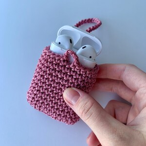 May include: A handmade, dusty rose crochet case for wireless earbuds. The case is open, revealing white earbuds inside. A matching crochet loop is attached for carrying. The case is held by a hand against a white background.