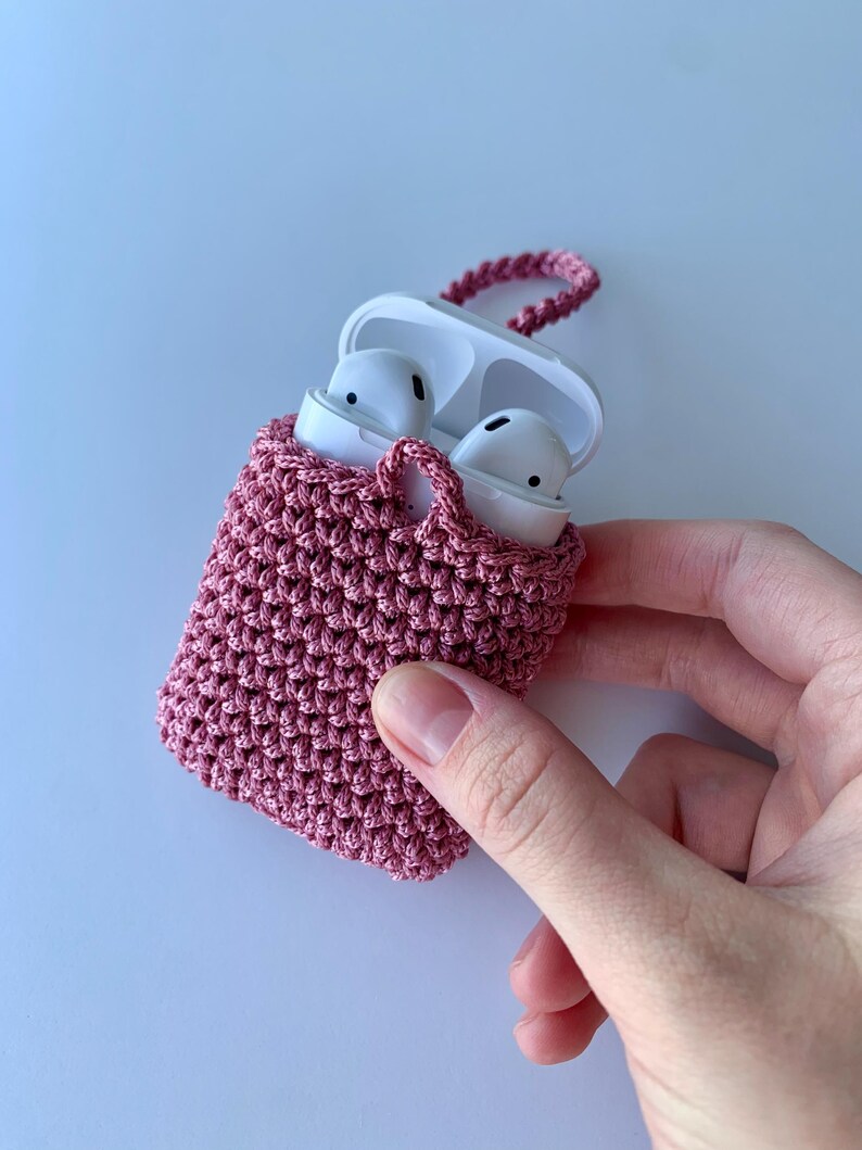 May include: A handmade, dusty rose crochet case for wireless earbuds. The case is open, revealing white earbuds inside. A matching crochet loop is attached for carrying. The case is held by a hand against a white background.
