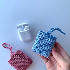 May include: A white AirPods case next to two crocheted cases, one pink and one blue. The cases are small, square, and have a loop at the top. The blue case is being held by a hand. The background is a light blue.