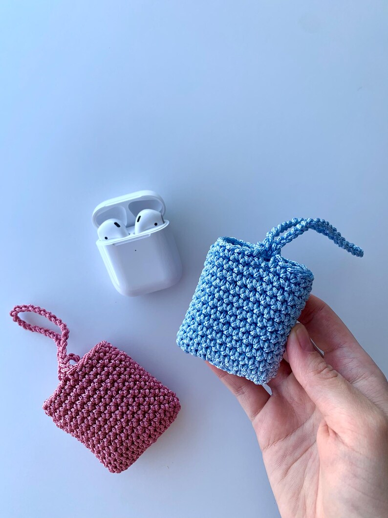 May include: A white AirPods case next to two crocheted cases, one pink and one blue. The cases are small, square, and have a loop at the top. The blue case is being held by a hand. The background is a light blue.