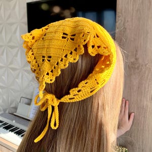 K&ouml;nnte beinhalten: Ein leuchtend gelbes, geh&auml;keltes Kopftuch mit einem gewellten Rand und dekorativen Aussparungen. Die Kopfbedeckung wird am Nacken mit einer passenden gelben Krawatte befestigt. Das Kopftuch ist handgefertigt und scheint aus Baumwollgarn zu bestehen.