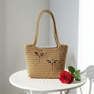Op de afbeelding: Een beige gehaakte tote bag met libellen uitsparingen. De tas heeft twee handvatten en staat op een witte tafel. Een rode roos met groene bladeren ligt naast de tas.