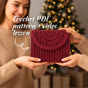 以下が含まれることがあります： 人が持っている、あずき色のクロシェクラッチ。フラップ開閉式で、リブ編みのボディと装飾的な半円形の上部が特徴です。画像には「Crochet PDF pattern + video lesson」と書かれています。