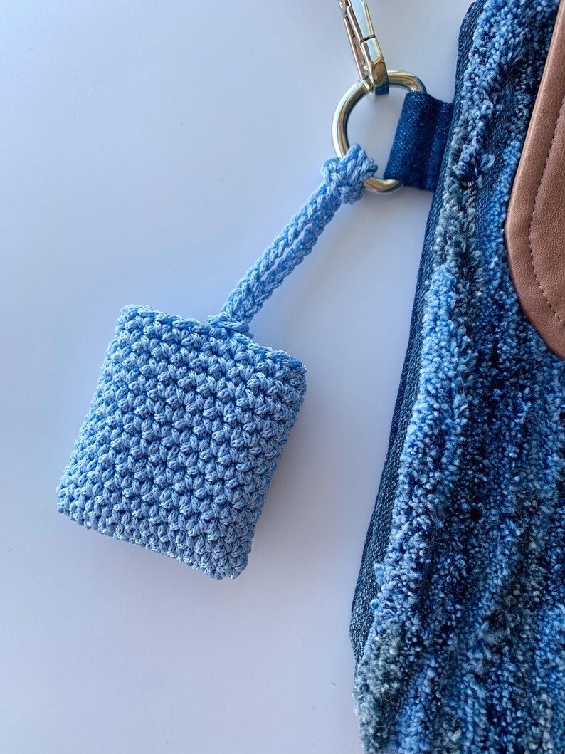 May include: A light blue, crocheted square pouch with a matching strap, attached to a silver metal ring. The pouch is connected to a denim bag with a brown leather accent.