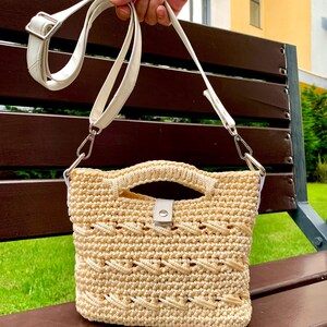 Puede incluir: Un bolso de mano de ganchillo beige con una correa y asa de cuero blanco. El bolso presenta un patr&oacute;n texturizado y un peque&ntilde;o cierre blanco. El bolso es sostenido por una mano.