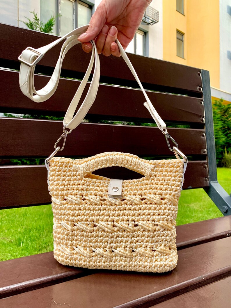 Puede incluir: Un bolso de mano de ganchillo beige con una correa y asa de cuero blanco. El bolso presenta un patr&oacute;n texturizado y un peque&ntilde;o cierre blanco. El bolso es sostenido por una mano.