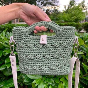 Puede incluir: Un bolso de mano de ganchillo verde salvia con un dise&ntilde;o trenzado. El bolso tiene un asa superior y una correa para el hombro desmontable. La correa y el cierre son de color crema claro. El bolso se sostiene frente a un fondo de hojas verdes.