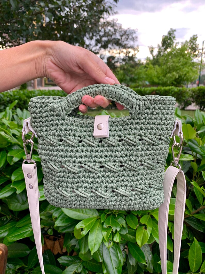 Puede incluir: Un bolso de mano de ganchillo verde salvia con un dise&ntilde;o trenzado. El bolso tiene un asa superior y una correa para el hombro desmontable. La correa y el cierre son de color crema claro. El bolso se sostiene frente a un fondo de hojas verdes.