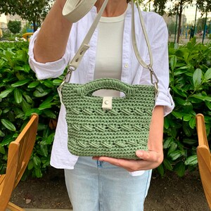 Puede incluir: Un bolso de mano verde tejido a ganchillo con una asa superior y una correa de hombro blanca desmontable. El bolso presenta un patr&oacute;n tejido y un cierre de cuero blanco. El bolso se muestra frente a un fondo de follaje verde.