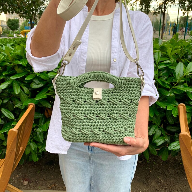 Puede incluir: Un bolso de mano verde tejido a ganchillo con una asa superior y una correa de hombro blanca desmontable. El bolso presenta un patr&oacute;n tejido y un cierre de cuero blanco. El bolso se muestra frente a un fondo de follaje verde.