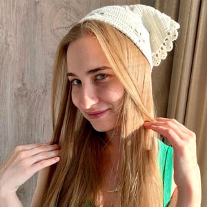 K&ouml;nnte beinhalten: Eine cremefarbene, geh&auml;kelte Kopfbedeckung mit einem gewellten Rand. Die Kopfbedeckung ist aus einem weichen, strukturierten Garn gefertigt und weist ein zartes Lochmuster auf. Der Artikel wird auf dem Kopf getragen.