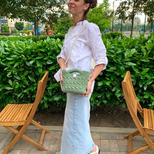 Puede incluir: Un peque&ntilde;o bolso verde tejido a ganchillo con una correa color crema. El bolso lo lleva una persona que viste una camisa blanca y una falda larga de mezclilla. El fondo presenta follaje verde y sillas de madera.