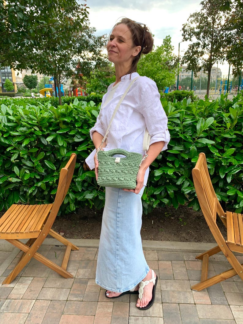 Puede incluir: Un peque&ntilde;o bolso verde tejido a ganchillo con una correa color crema. El bolso lo lleva una persona que viste una camisa blanca y una falda larga de mezclilla. El fondo presenta follaje verde y sillas de madera.