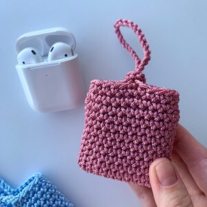 May include: A pink crocheted case for wireless earbuds, held by a hand. The case is square with a loop at the top. A white earbud charging case and a blue crocheted case are also visible.