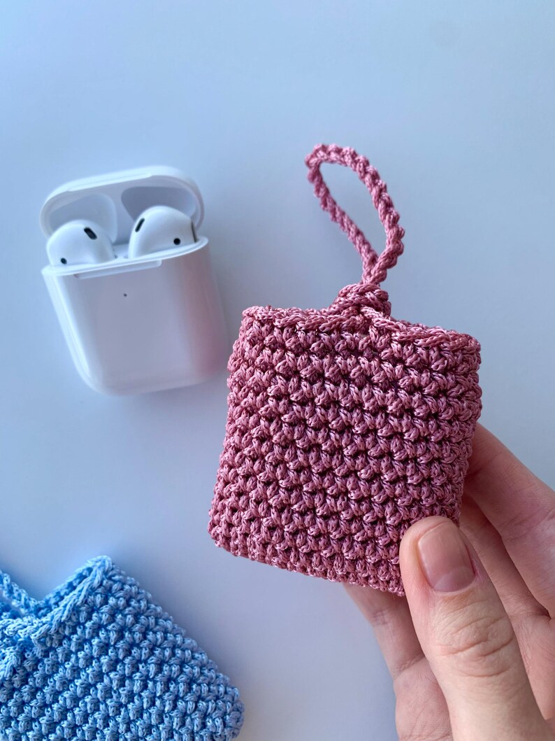 May include: A pink crocheted case for wireless earbuds, held by a hand. The case is square with a loop at the top. A white earbud charging case and a blue crocheted case are also visible.