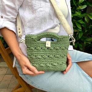 Puede incluir: Un bolso de mano verde tejido a ganchillo con una correa y asa color crema. El bolso presenta un patr&oacute;n texturizado y un peque&ntilde;o cierre de solapa con un bot&oacute;n. La correa es ajustable y se sujeta al bolso con cierres plateados.