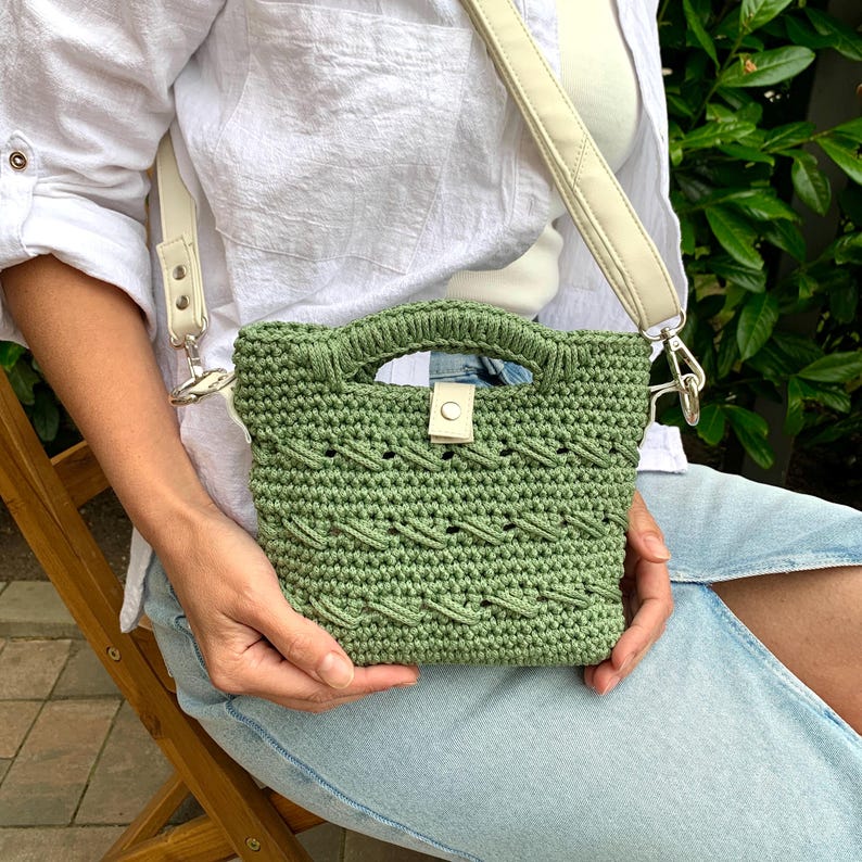 Puede incluir: Un bolso de mano verde tejido a ganchillo con una correa y asa color crema. El bolso presenta un patr&oacute;n texturizado y un peque&ntilde;o cierre de solapa con un bot&oacute;n. La correa es ajustable y se sujeta al bolso con cierres plateados.