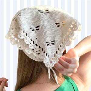 K&ouml;nnte beinhalten: Cremefarbene H&auml;kelkopfbedeckung mit bogenf&ouml;rmigen Aussparungen. Die Kopfbedeckung hat einen gewellten Rand und Bindeb&auml;nder. Die Kopfbedeckung ist aus einem weichen, strukturierten Garn gefertigt.