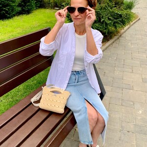 Puede incluir: Una mujer con gafas de sol, camisa blanca y falda vaquera est&aacute; sentada en un banco del parque. Un peque&ntilde;o bolso beige tejido con correa para el hombro descansa en el banco. Lleva sandalias blancas.