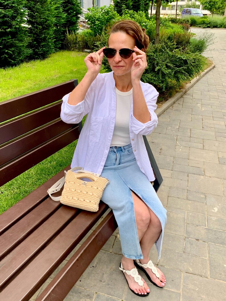 Puede incluir: Una mujer con gafas de sol, camisa blanca y falda vaquera est&aacute; sentada en un banco del parque. Un peque&ntilde;o bolso beige tejido con correa para el hombro descansa en el banco. Lleva sandalias blancas.