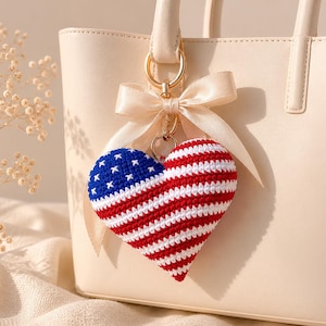 May include: A crocheted heart-shaped keychain with a design resembling the American flag. The keychain is red, white, and blue, with a blue section featuring white stars and red and white stripes. It hangs from a beige handbag.