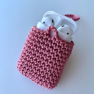 May include: A pink crocheted case for wireless earbuds. The case has a handle and is open, revealing white earbuds inside. The case is made of a textured, woven material. The background is a light blue.