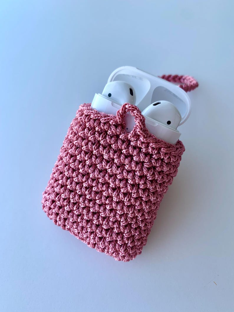 May include: A pink crocheted case for wireless earbuds. The case has a handle and is open, revealing white earbuds inside. The case is made of a textured, woven material. The background is a light blue.