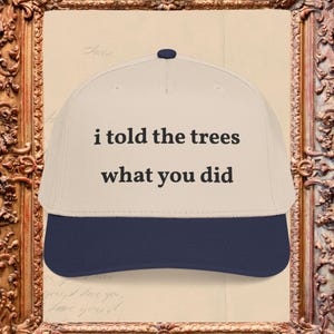 May include: A beige baseball cap with a dark blue brim and a dark blue button on top. The front of the cap features the black text "i told the trees what you did" printed in a simple, sans-serif font across the center of the crown.