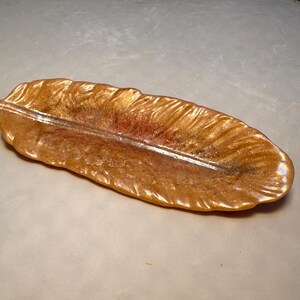 May include: A gold feather-shaped trinket dish with a metallic sheen. The dish is made of resin and has a detailed feather design.