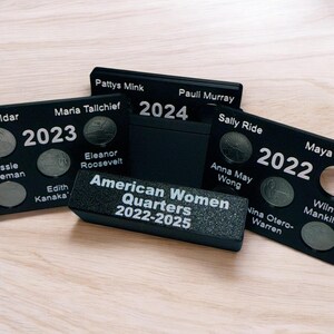 Pode incluir: Painéis de exibição pretos para os American Women Quarters, com ranhuras para moedas e nomes como Sally Ride e Maria Tallchief. Os painéis estão rotulados com os anos 2022, 2023 e 2024, e o texto "American Women Quarters 2022-2025".